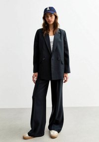 Blazer nero a righe con doppi bottoni, abbinato a pantaloni larghi coordinati. Indossato sopra una maglietta bianca, completato da un berretto blu navy.