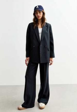 Black pinstripe blazer with double buttons, paired with matching wide-leg trousers. Worn over a white top, complemented by a navy cap.