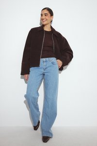 Giacca di velluto a coste marrone con chiusura a zip, abbinata a jeans a gamba dritta blu chiaro e tacchi a punta marrone scuro. Texture liscia.