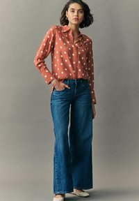 Rustfarbiges Langarmshirt mit weißen Pünktchen; kombiniert mit hoch taillierten, ausgestellten blauen Jeans; das Model steht vor einem neutralen Hintergrund.