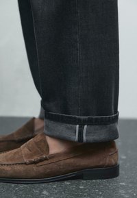 Mocassini in suede marrone con dettagli di cucitura, abbinati a jeans di denim scuro arrotolati con un orlo in un tono più scuro.