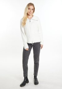 Maglione bianco con zip, caratterizzato da un motivo a treccia e una texture a coste. Abbinato a pantaloni grigi a vestibilità aderente e stivaletti neri alla caviglia.