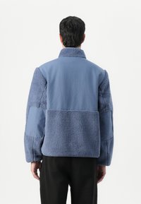 Giacca in pile blu con un tessuto testurizzato e morbido e un pannello in tessuto liscio. Presenta un collo alto e una vestibilità ampia, con maniche lunghe.