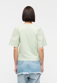 Γυναίκα με κοντά καστανά μαλλιά, φορώντας ανοιχτό πράσινο τοπ με φουσκωτά μανίκια και σκισμένη τζιν φούστα πάνω από φαρδιά τζιν, στέκεται μπροστά σε λευκό φόντο.