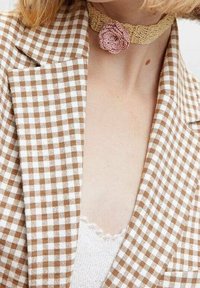 Blazer en vichy marron et blanc avec un col cranté, associé à un collier tour de cou en dentelle beige orné d'une fleur en crochet rose.