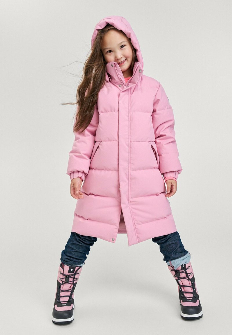 Abrigo tipo puff rosa con capucha, que presenta un cierre de presión en el frente y puños elásticos. Combinado con botas de invierno negras y rosas. Lleva unos vaqueros azules con puños.