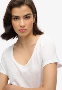 Camiseta blanca de manga corta con cuello en V, hecha de tejido texturizado. Presenta un discreto parche con el logo en el lado izquierdo del pecho. Diseño simple, ajuste relajado.