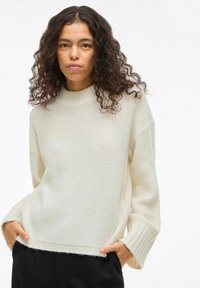 Kreemikas värvi kudumülikond kõrge ribiga kraega, laiade varrukate ja lõdva lõikega. Omab tekstuurset mustrit ja külgmist lõhikut ääres.