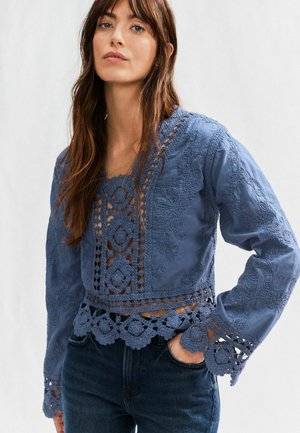 Blå broderet bluse med lange ærmer, hæklede detaljer, bølget kant og udsparinger, parret med mørke denim jeans.