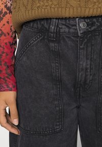 Black denim pants with two large side pockets, textured fabric, high waist, and visible stitching details. Worn with a patterned long-sleeve top.