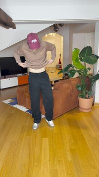 Καφέ μακριά μανίκια πουκάμισο, μαύρο παντελόνι και ασημένια παπούτσια; προσαρμόζοντας τα ρούχα ενώ στέκεται σε ένα δωμάτιο με καφέ καναπέ και φυτό.