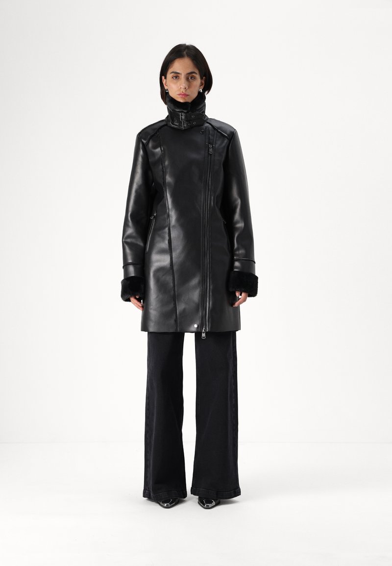 Cappotto in ecopelle nera con colletto alto, chiusura con zip e polsini foderati in pelliccia. Abbinato a jeans neri a gamba larga e scarpe con tacco.