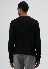 Pull noir côtelé à manches longues, avec un col rond et des coutures visibles. Porté avec un pantalon gris clair, mettant en valeur un tissu texturé.