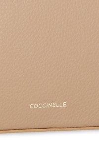 Pelle testurizzata beige con una finitura liscia, caratterizzata da un logo dorato in rilievo "COCCINELLE" nella parte inferiore. Design rettangolare piatto.