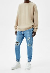 Sudadera beige con cuello redondo, combinada con jeans claros desgastados que tienen detalles rasgados, y zapatillas blancas con cordones texturizados.