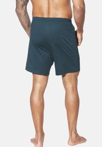 Oben ohne Mann mit tätowiertem linken Arm, der dunkelblaue elastische Shorts trägt und barfuß vor einem schlichten weißen Hintergrund steht.