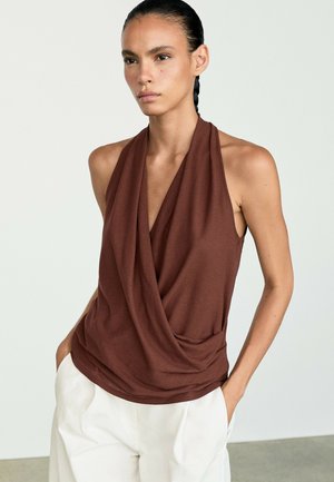 Femme aux cheveux foncés tressés, portant un haut marron drapé sans manches et un pantalon blanc, debout les mains dans les poches devant un fond uni.