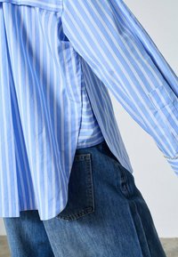 Lyseblå skjorte med striber, lange ærmer og løs pasform, kombineret med højtaljede denimbukser. Har en sidesprække og opsmøgede ærmer.