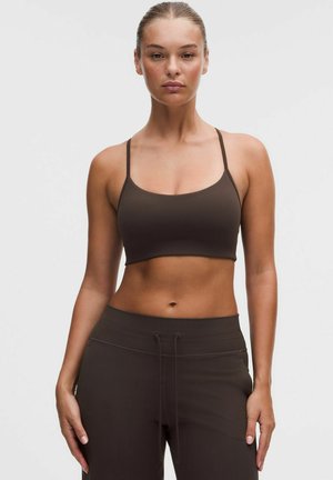 Kvinde stående oprejst iført en mørkebrun cropped tanktop og matchende højttaljede bukser mod en ensartet lys baggrund.