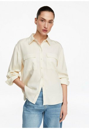 Frau mit zurückgekämmtem Haar, die ein cremefarbenes Hemd mit hochgekrempelten Ärmeln und hellblaue Jeans trägt, steht mit einer Hand in der Tasche.