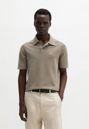 Homme portant un polo beige à manches courtes rentré dans un pantalon beige clair avec une ceinture noire, debout devant un fond blanc uni.