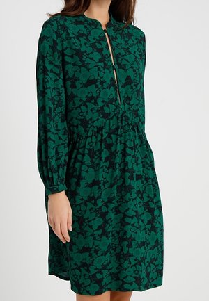 Nainen pukeutuneena pitkähihainen, vihreä ja musta kukkakuvioinen polvipituinen mekko, jossa on kerrostettu vyötärö ja pieni avonainen aukko edessä kaula-aukossa.