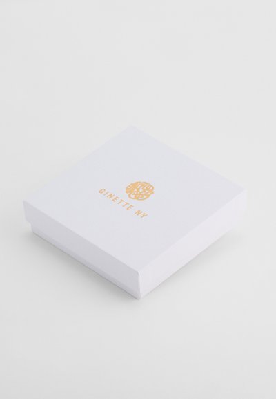 Balts kvadrāta formas rotaslietu kastē ar zelta logotipu un tekstu "GINETTE NY" uz vāka uz vienkārša baltā fona.