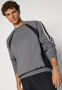 Sudadera gris con paneles laterales negros y ribete blanco. Presenta puños acanalados y un pequeño logo en el pecho. Tejido suave y texturizado.
