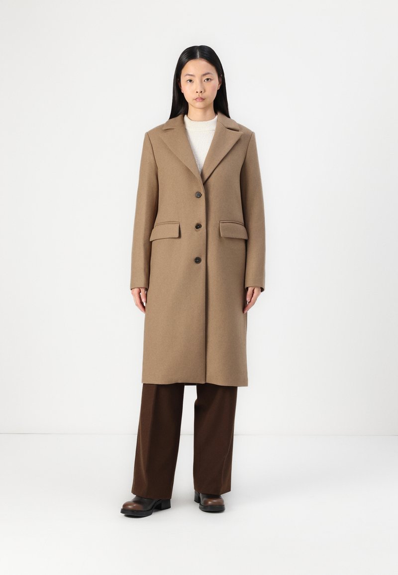 Cappotto di lana marrone con colletto a punta, tre bottoni e due tasche frontali, sopra un maglione crema e pantaloni marroni.