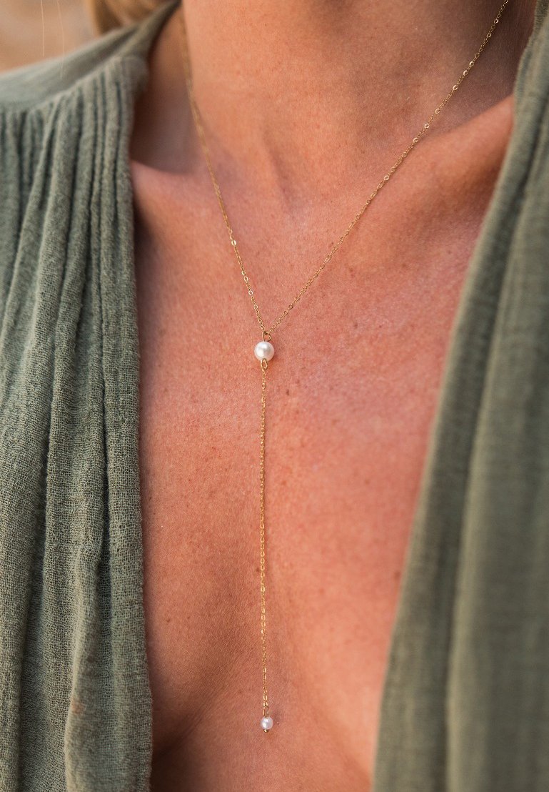 Collana delicata in oro con due piccole perle bianche indossata sul petto nudo di una persona, insieme a un indumento verde dalla texture.