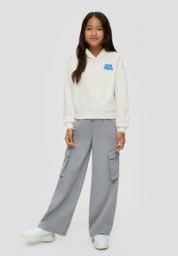 Weiße Kapuzenjacke mit blauem "lost vibes"-Schriftzug, kombiniert mit lockeren grauen Cargo-Hosen mit großen Seitentaschen und weißen Sneakers.