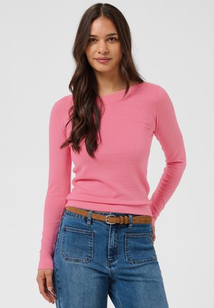 Femme aux longs cheveux foncés portant un haut rose à manches longues, un jean bleu avec de grandes poches avant et une ceinture marron, debout devant un fond uni.