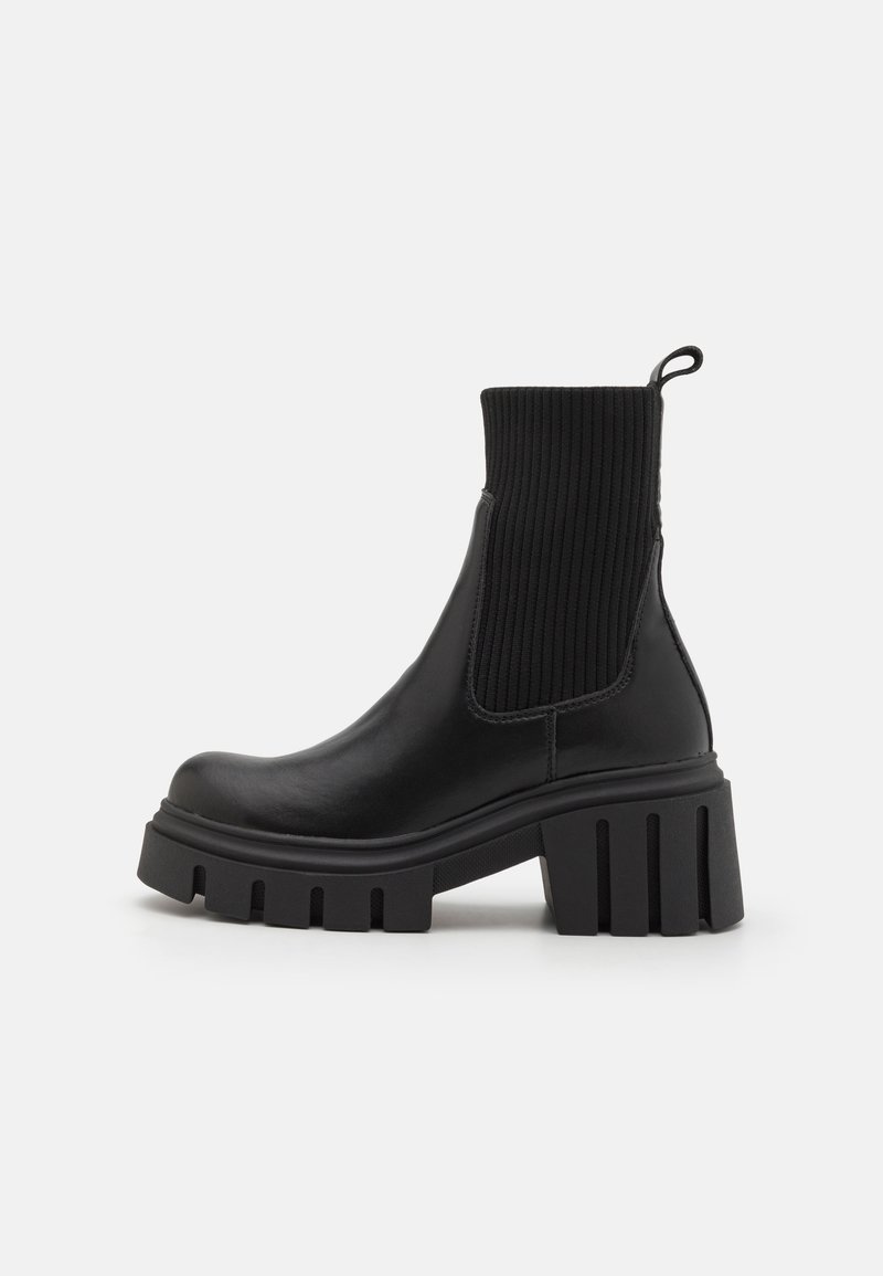 Bottine noire à talon carré épais avec tige en tricot côtelé, languette au talon et semelle crantée épaisse sur fond blanc.
