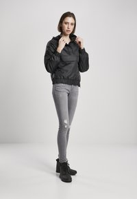Črna najlon kapuca z elastičnimi manšetami in pasom, ki vključuje sprednji žep in nastavljivo kapuco z vrvico, oblečena z svetlo sivo uničenimi jeans hlačami.