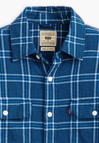 Chemise en flanelle à carreaux bleus avec un motif de checks blancs, col boutonné, deux boutons à l'avant et poche sur la poitrine gauche. Détails de l'étiquette visibles.