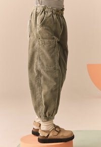 Grüne Cordhose mit lockerer Passform, elastischem Bund, seitlichen Taschen und gerippter Textur, kombiniert mit beigen Schnallenschuhen und weißen Socken.