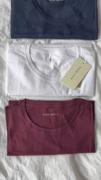 Τρία διπλωμένα T-shirt σε ναυτικό μπλε, λευκό και μπορντό βαμβάκι, με στρογγυλές λαιμόκοψεις και εκτυπωμένα ετικέτες μάρκας. Μια ετικέτα είναι προσαρτημένη στο λευκό πουκάμισο.