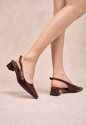 Pieds portant des escarpins slingback en cuir marron foncé avec des talons blocs bas sur un sol et un fond texturés clairs.