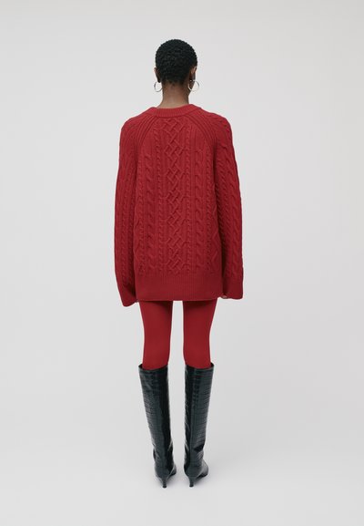 Asmuo atsukęs nugarą, vilkintis raudoną megztinį su akyčių raštu, atitinkamas raudonas leggingas, juodas aukštas tekstūruotas batus iki kelių ir sidabrines auskarių žiedus, ant vientiso fono.