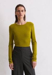Langärmliges, olivgrünes Oberteil mit Rundhalsausschnitt, kombiniert mit hoch sitzenden, dunkelgrauen, gestreiften Hosen. Weiche Stofftextur sichtbar.