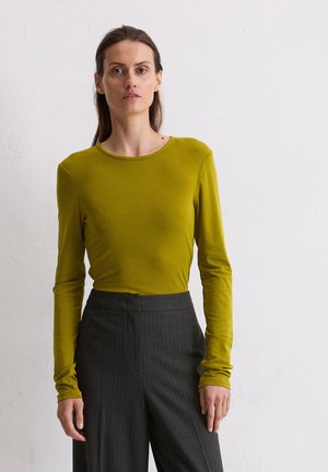 Haut à manches longues, vert olive, avec un col rond, associé à un pantalon taille haute à rayures noires et grises. Texture du tissu douce et visible.