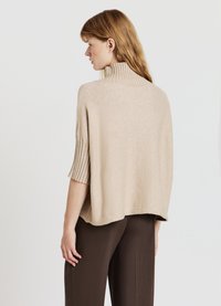 Beige Strickpullover mit hohem, geripptem Kragen und kurzen, ausgestellt geschnittenen Ärmeln. Der Rücken ist etwas länger und hat eine glatte, weiche Textur.