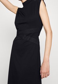 Černé šaty s vysokým výstřihem, krátkými rukávy a nařasením. Obsahují logo na pásku a mají přiléhavý střih. Hladká textura.