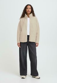 Beežist fliisjope kõrge kraega, täieliku lukuga ja küljetaskutega; kombineeritud tumedate laia säärega teksade ja beežide spordijalatsitega. Tekstuur on pehme ja hubane.