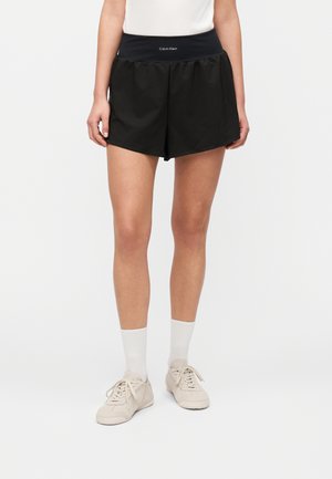 Μαύρα σορτς με λεία υφή και φαρδιά μέση που φέρει το λογότυπο Calvin Klein. Φοριούνται με λευκές κάλτσες αστραγάλου και ανοιχτό μπεζ παπούτσια.