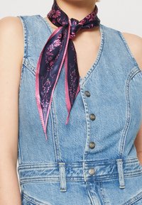 Jeansoverall i blått tyg, med V-ringning, metallknappar och en blommönstrad mörkblå och rosa silkes scarf knuten runt halsen.