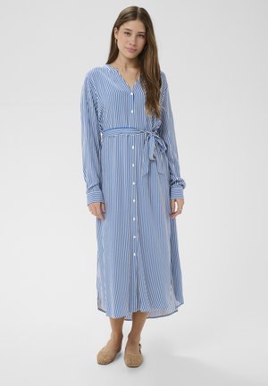 Naine, kes kannab sinise ja valge vertikaalsete triipudega nööbitavat kleiti, mis on vöökohalt seotud, ning beeže kootud tasaseid jalanõusid, seisab ühevärvilise tausta ees.