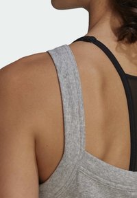 Haut de sport gris sans manches avec une finition texturée, doté de larges bretelles et superposé à un soutien-gorge de sport noir en dessous.