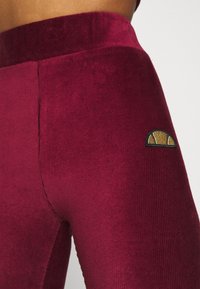 Burgundin samettihousut, joissa on sileä pinta, korkea vyötärö ja pieni kultaketjulla varustettu logo reidessä.