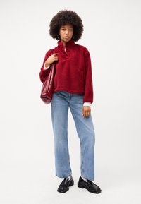 Rød fleece genser med halv-zip hals, frontlomme, matchende lyseblå vide jeans og svarte loafers, holder en kastanjefarget totebag.